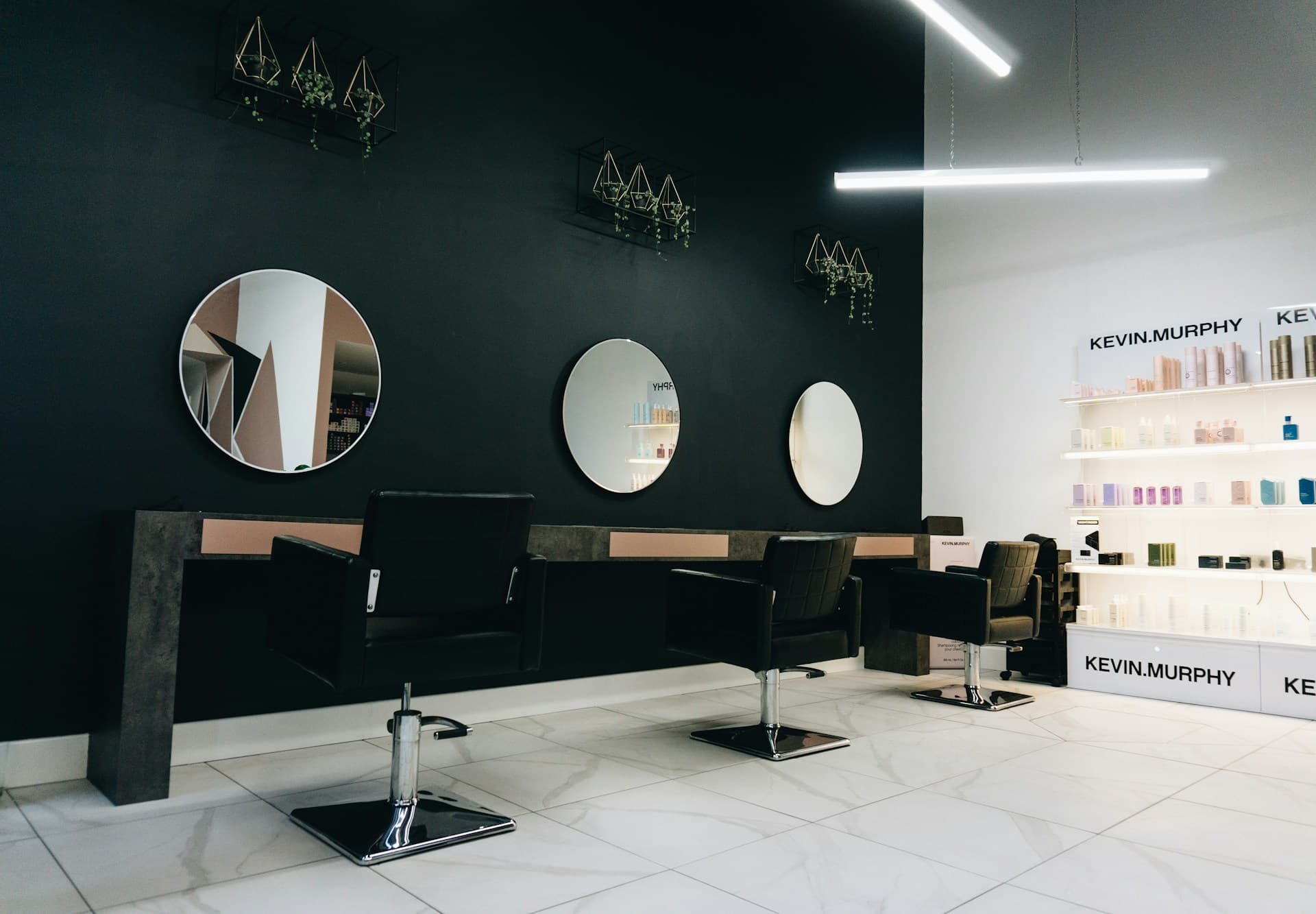 Salón de peluquería con sillones negros, espejos redondos y una estantería de productos
